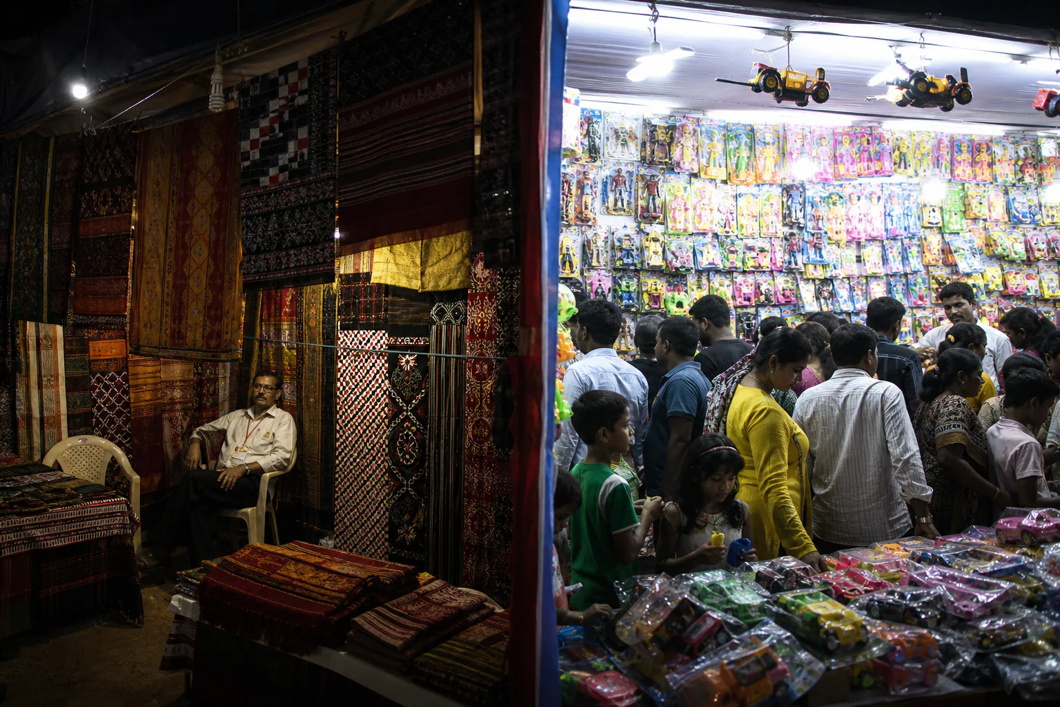 A traditional textile stall lit warmly beside a crowded stall of brightly packaged plastic toys